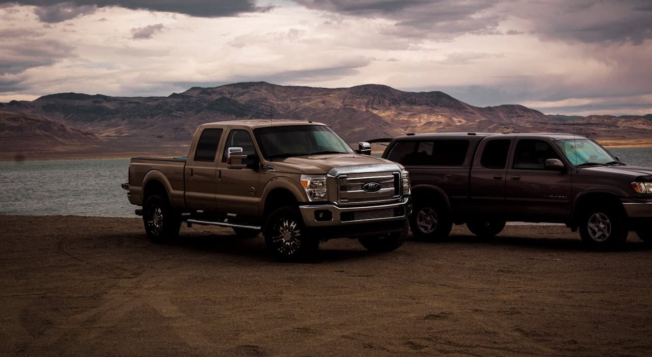 Electric pickup truck Thailand: Two pickup trucks parked on a sandy lakeshore, with mountains and cloudy skies in the background.