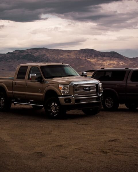 Electric pickup truck Thailand: Two pickup trucks parked on a sandy lakeshore, with mountains and cloudy skies in the background.